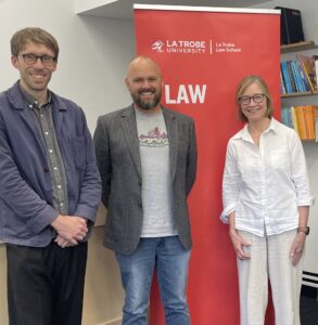 Piers, Chris, and Jill standing smiling in front of the La Trobe Law School banner